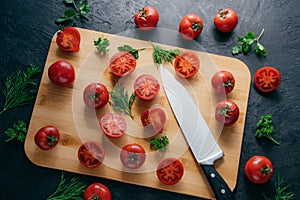 Tasty ripe tomatoes sliced on chopping Board with greenery, sharp knife near, isolated over dark background. Kitchenware. Top view