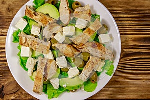 Tasty salad with fried chicken breast, green olives, feta cheese, avocado, lettuce and olive oil on wooden table. Top view