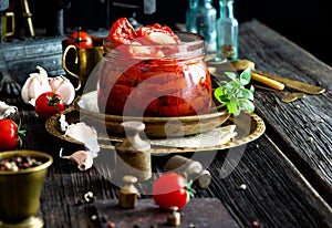 Tasty sun dried tomatoes with garlic, pepper, basil, olive oil in glass jar stands on brass plates on rustic wooden table