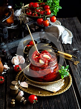 Tasty sun dried tomatoes with garlic, pepper, basil, olive oil in glass jar stands on brass plates on rustic wooden table