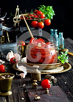 Tasty sun dried tomatoes with garlic, pepper, basil, olive oil in glass jar stands on brass plates on rustic wooden table