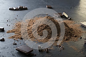 Tasty sweet pieces of chocolate and heap of cocoa powder on the dark table, close up