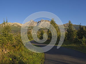 Tatra Mountains range with asphalt road curve, golden hour light, way from Tatranska Lomnice to Start with green trees and grass.