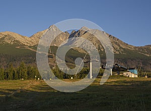 Tatra Mountains range gold colored sunrise light , ski lift at Tatranska Lomnice with green trees and grass. Early autumn panorama