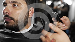 Tattoed barber shears the customer`s beard by using trimmer at the barber shop, man`s haircut and shaving at the