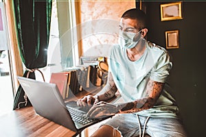 Tattoo man freelancer in face mask, using laptop for remotely working online in cafe