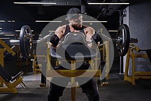 Tattooed athlete in black sport gloves, shorts, vest, cap. Lifting barbell, training his biceps, sitting on preacher