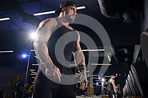 Tattooed male in black shorts, vest, cap. Going to do exercises with dumbbells, looking at mirror near set of black