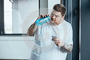 Tattooed man using smartphone and drinking water from sport bottle at gym