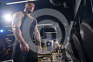 Tattooed, muscular bearded guy in black shorts, vest, cap. Looking at you, standing near set of black dumbbells. Dark