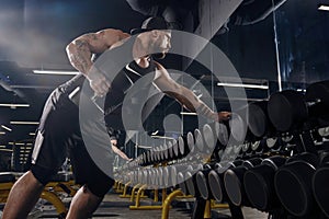 Tattooed, muscular guy in black shorts, vest and cap. Exercising with dumbbells for training his triceps in dark gym