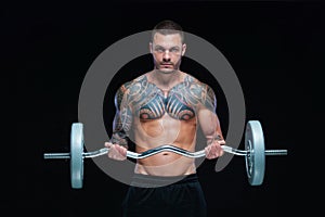 Tattooed strong muscular athletic man pumping up muscles with barbell on black background