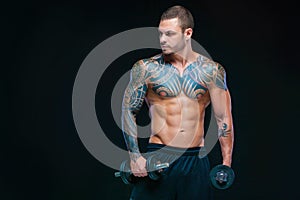 Tattooed strong muscular athletic man pumping up muscles with dumbbells on black background