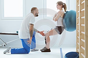 Tattooed trainer assisting a girl during physiotherapy exercises with ball