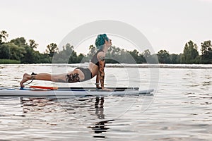 tattooed woman with blue hair practicing yoga on paddleboard in water. Upward facing dog pose