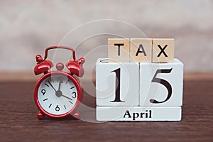 Tax with 15th April. Alphabet and calendar wooden block cube with number and Red alarm clock. On table dark plank wooden