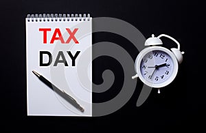 TAX DAY is written in a white notepad near a white alarm clock on a black background