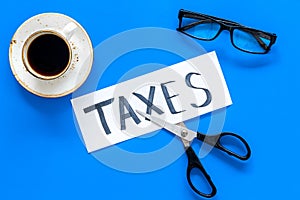 Tax reduce concept. Sciccors and paper with word Taxes on blue background with coffee and glasses top view