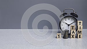 Tax time concept. An alarm clock with stack of money and wooden block with text on grey background with copy space
