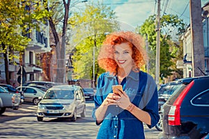 Taxi app. Pretty happy smiling urban woman using smart phone, standing on city street on sunny day waiting for taxi. Beautiful