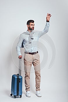 Taxi. Young and handsome man with a luggage is raising his hand while standing in studio on a grey background. Full