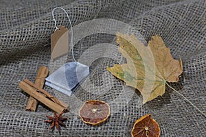 Tea bag, dried maple leaves on a rough linen cloth. Nearby are cinnamon, dried orange slices and anise. Autumn still life