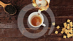 Tea being poured into glass tea cup on him located brown sugar and spoon with tea leaves on brown wooden table