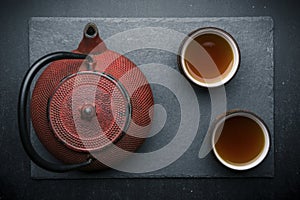 Tea composition with ceramic tea cups and red iron teapot on dark stone background