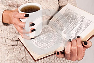 Tea cup and book in young women hands with black nails and sweater