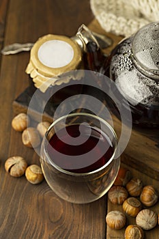 Tea in glass cup with teapot, jam in jar and knitted blanket near, with hazelnut at wood background, with spoon near.