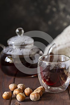 Tea in glass cup with teapot and knitted blanket near, with hazelnut at wood background.