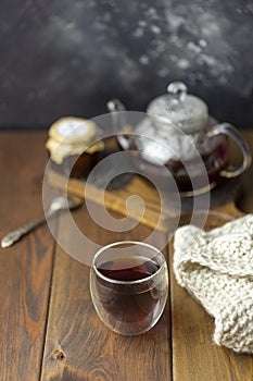 Tea in glass cup with teapot and knitted blanket near, with jam in jar at wood background, with spoon near.