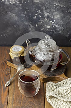 Tea in glass cup with teapot and knitted blanket near, with jam in jar at wood background, with spoon near.