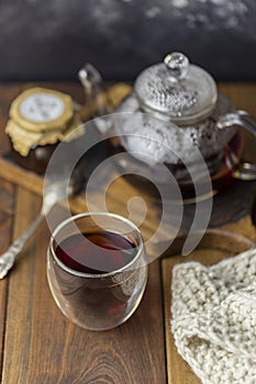 Tea in glass cup with teapot and knitted blanket near, with jam in jar at wood background, with spoon near.