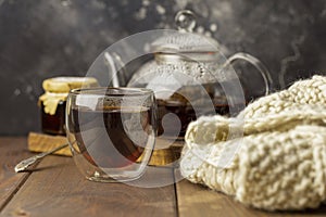 Tea in glass cup with teapot and knitted blanket near, with jam in jar at wood background, with spoon near.