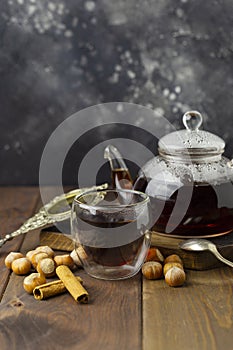 Tea in glass cup with teapot and knitted blanket near, with vanilla pod and hazelnut at wood background, with spoon and strainer n