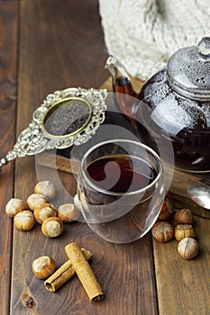 Tea in glass cup with teapot and knitted blanket near, with vanilla pod and hazelnut at wood background, with spoon and strainer n