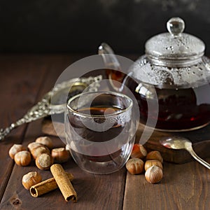 Tea in glass cup with teapot near, vanilla pod and hazelnut at wood background, with spoon and strainer near.