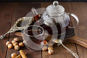 Tea in glass cup with teapot near, vanilla pod and hazelnut at wood background, with spoon and strainer near.