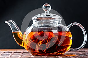 Tea in a glass teapot with a blooming large flower. Teapot with exotic green tea-balls blooms flower. Tea ceremony on a dark