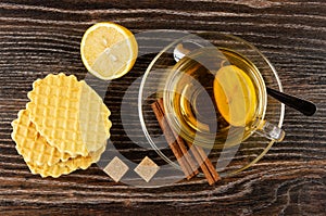 Tea with lemon in cup, cinnamon, spoon on saucer, wafers, sugar, half of lemon on wooden table. Top view