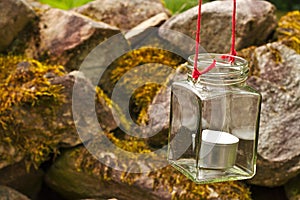 tea light candle ind glass hanging from a tree on a red rope with blurred stones in the background