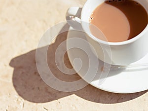 Tea with milk in a white cup in the morning sun with harsh shadows on a textured wood surface. Warm tones.