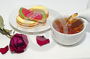 Tea is poured from a teapot into a cup, various cookies and a dried rose on a white background. Selective focus