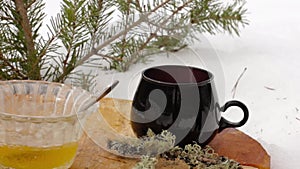 Tea set with honey: pouring hot tea from transparent teapot to black cup on a wooden stand among the snow