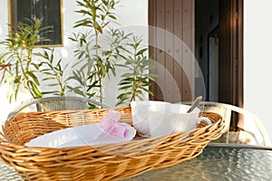 tea set in wicker tray on glass table in sunny garden, wooden door to house open, tea drinking on terrace with green plants,