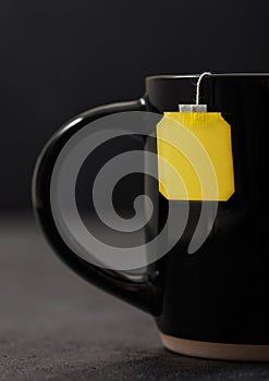 Teabag with yellow blank tag of black tea in black ceramic cup on dark board