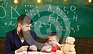 Teacher with beard, father teaches little son in classroom, chalkboard on background. Boy, child on calm face holds