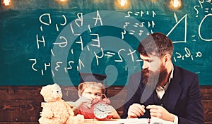 Teacher with beard, father teaches little son in classroom, chalkboard on background. Boy, child on calm face holds