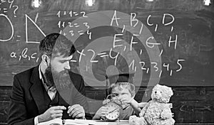 Teacher with beard, father teaches little son in classroom, chalkboard on background. Boy, child on calm face holds